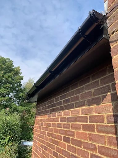 Black gutter and downpipe installed on red brick building exterior with green foliage visible