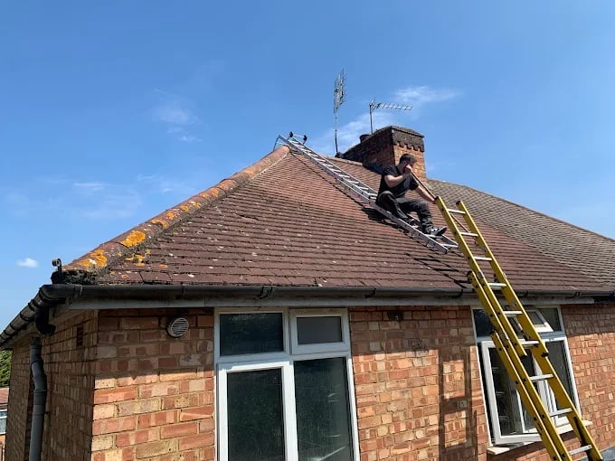 Roofer on ladder working on brown tiled roof of brick bungalow against blue sky