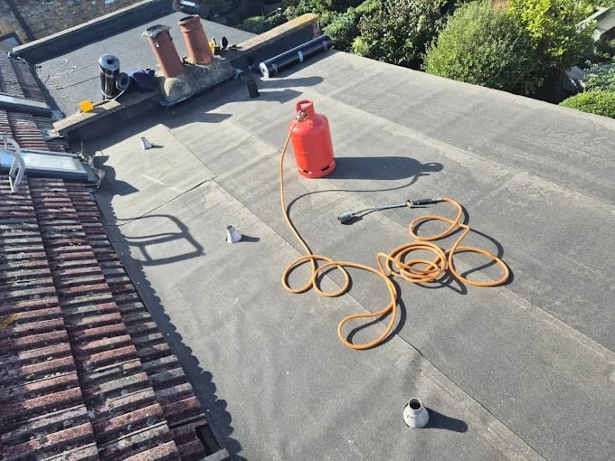 Roofer in red shirt standing on flat roof with coiled orange hoses and equipment during roof maintenance work